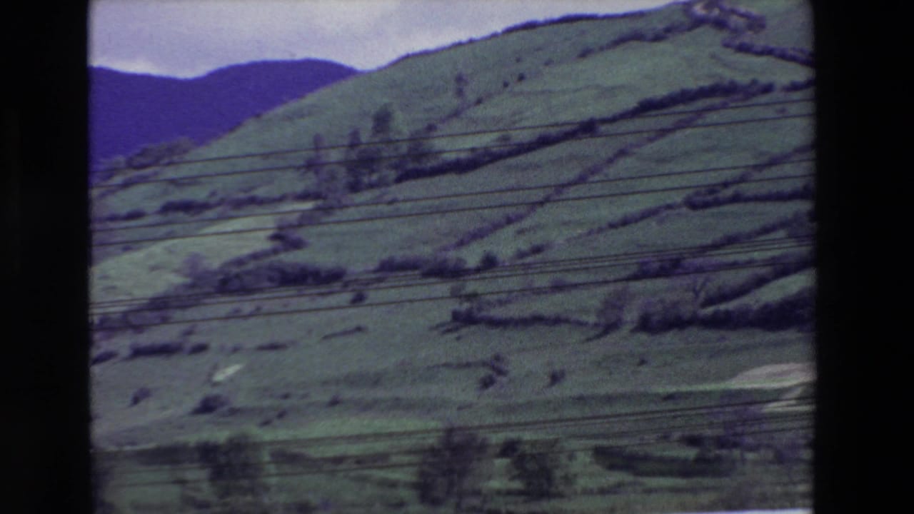 1969 8mm frame of a green rural hillside in Spain, with terraced field lines, dark distant hills, and overhead utility wires crossing the view.