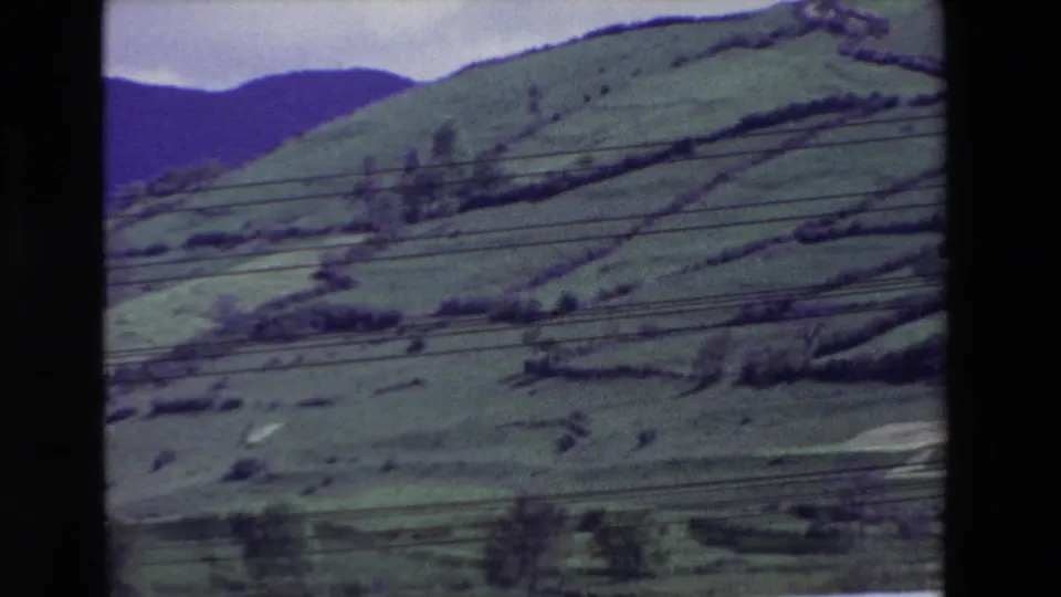 1969 8mm frame of a green rural hillside in Spain, with terraced field lines, dark distant hills, and overhead utility wires crossing the view.