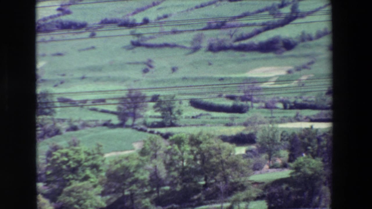 The pan reaches a rural valley with green fields, hedgerows, scattered trees, and utility lines visible across the 8mm frame.