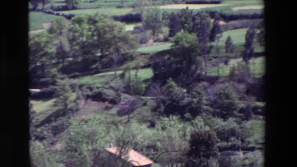 Rural valley detail from the 1969 home-movie pan, showing trees, hedgerows, grassy fields, and the edge of a tile-roofed building near the bottom of frame.