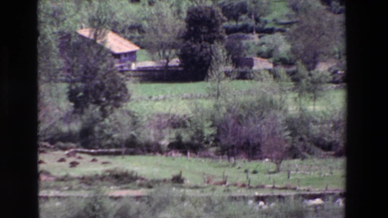 1969 8mm view in Spain of a rural farmhouse set beyond green fields, trees, shrubs, and low walls.
