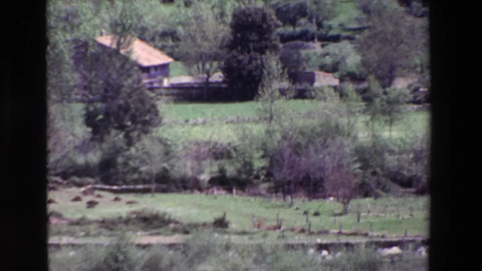 1969 8mm view in Spain of a rural farmhouse set beyond green fields, trees, shrubs, and low walls.