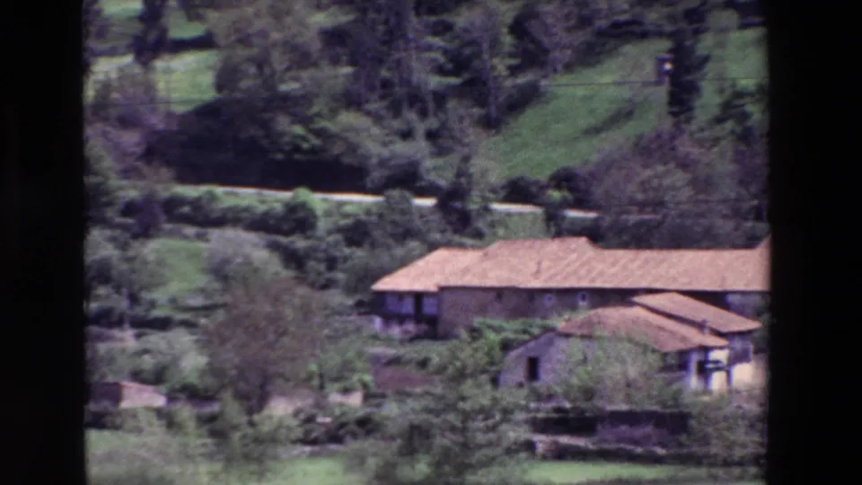 Distant 1969 view of rural buildings in Spain with terracotta tile roofs, trees, shrubs, a stone wall, and green hillside terrain.