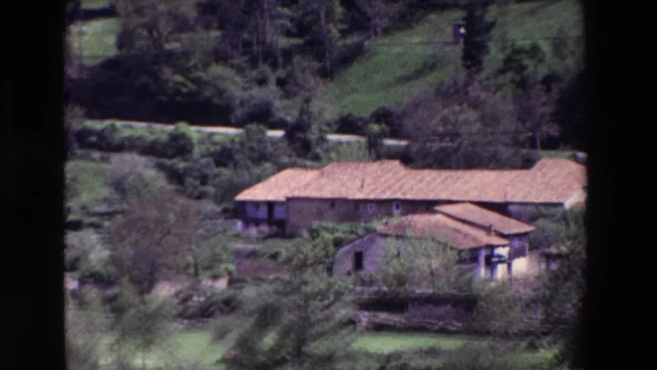 1969 8mm home-movie frame of a rural building with terracotta roof set in green hillside vegetation, with dark film borders visible.