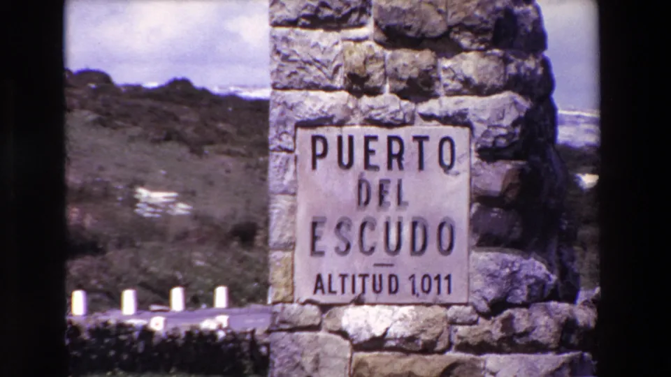 Close roadside view of a stone monument sign reading Puerto del Escudo Altitud 1,011, with rocky ground and pale roadside posts behind it.
