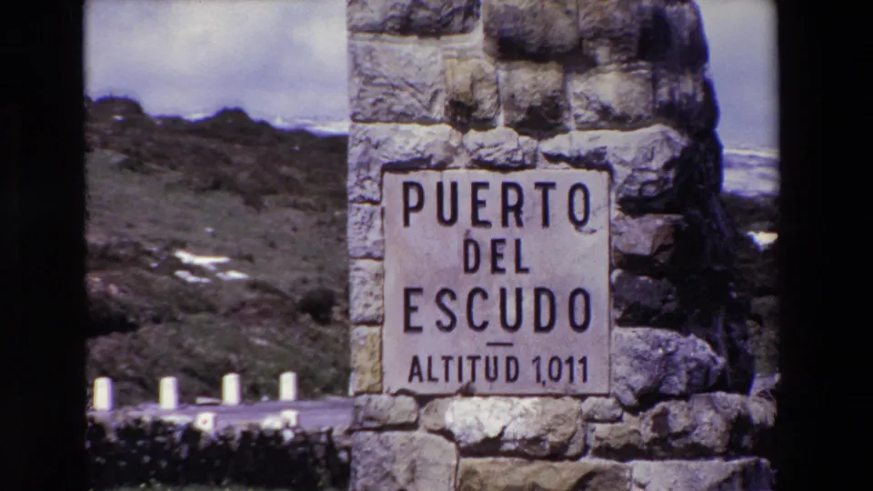 8mm frame of the Puerto del Escudo Altitud 1,011 stone sign, framed close with dark film borders and mountainous roadside terrain in the background.
