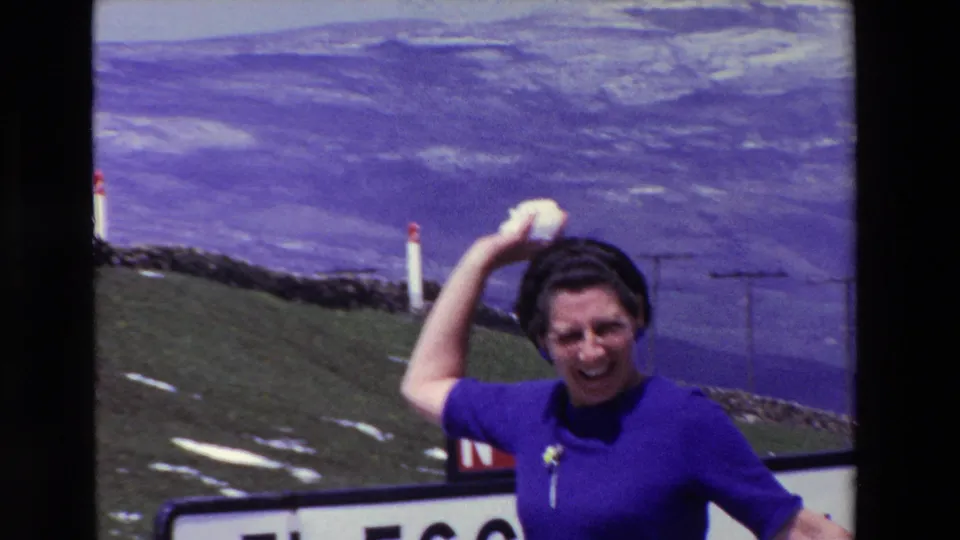 A woman in a purple dress raises her arm and smiles near N-623 and El Escudo signage at a Spanish mountain pass, with green slopes and a purple-blue distant horizon behind her.