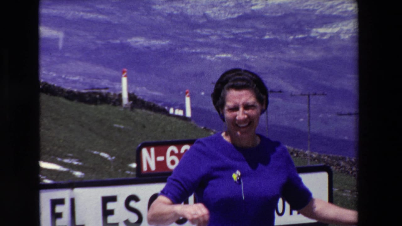 Woman smiles toward the camera beside road signs and green mountain slopes.