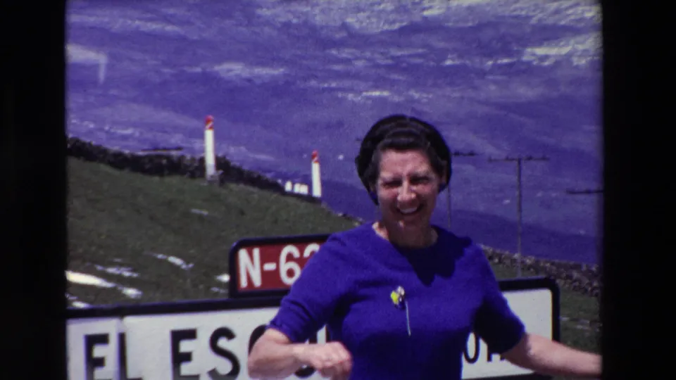 The woman faces the camera smiling beside the partially visible El Escudo and N-623 signs, with grassy uplands, pale patches on the slope, and utility poles in the background.