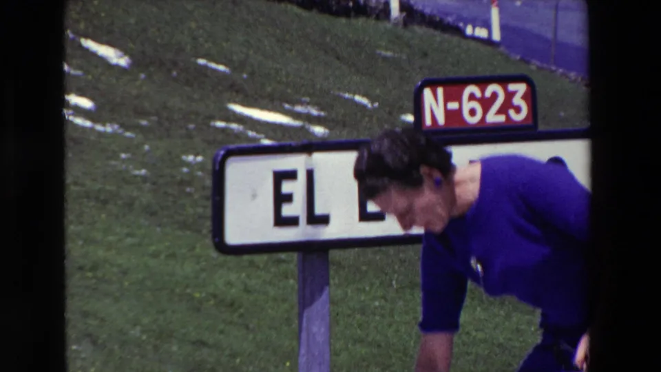 The woman bends forward playfully in front of the El Escudo road sign, with the N-623 marker, green hillside, and small pale ground patches visible behind her.