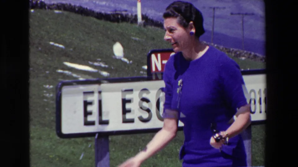 The woman appears in profile near the El Escudo sign, holding a small dark object as she gestures, with the red N-623 marker and grassy mountain slope behind her.