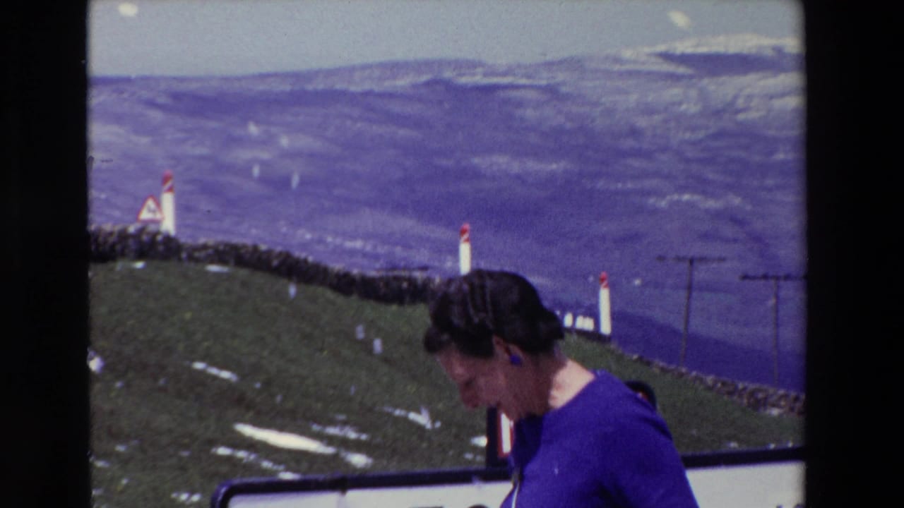 A woman in a purple dress stands near a roadside at Puerto del Escudo, Spain, with white posts, snow patches, and distant mountain slopes visible in 1969 8mm footage.
