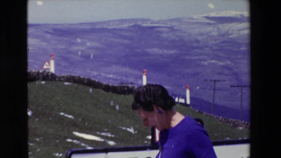 A woman in a purple dress stands near a roadside at Puerto del Escudo, Spain, with white posts, snow patches, and distant mountain slopes visible in 1969 8mm footage.