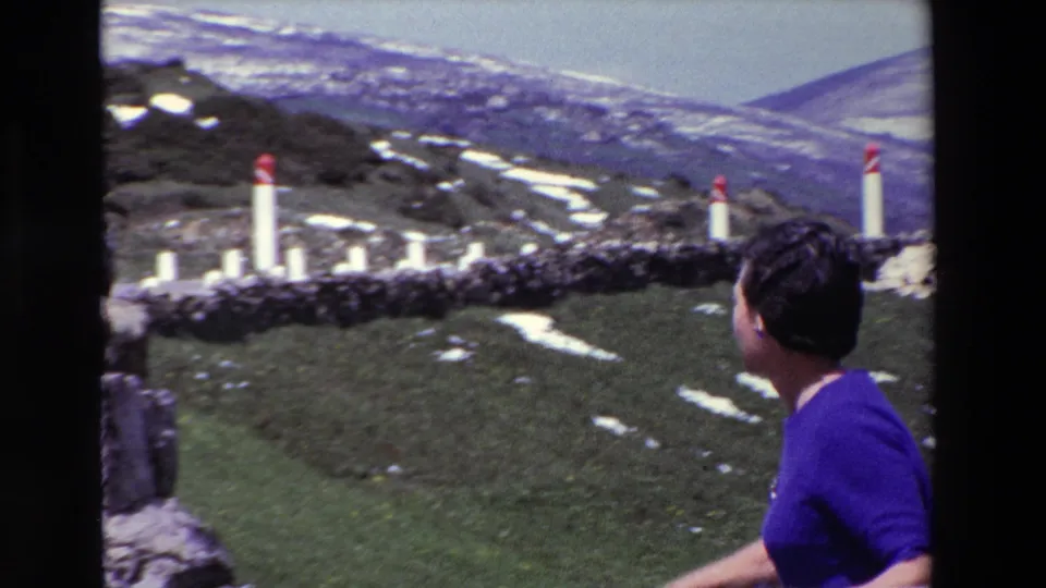 The woman turns toward the snow-dusted hillside at Puerto del Escudo, with red-topped white posts, stone edging, and layered mountain terrain in the background.