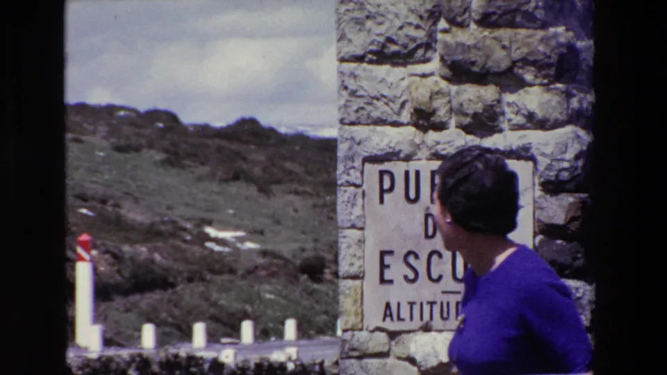 A woman in a purple dress stands beside a stone wall and partially visible Puerto del Escudo altitude sign, with roadside posts and rocky mountain ground behind her.