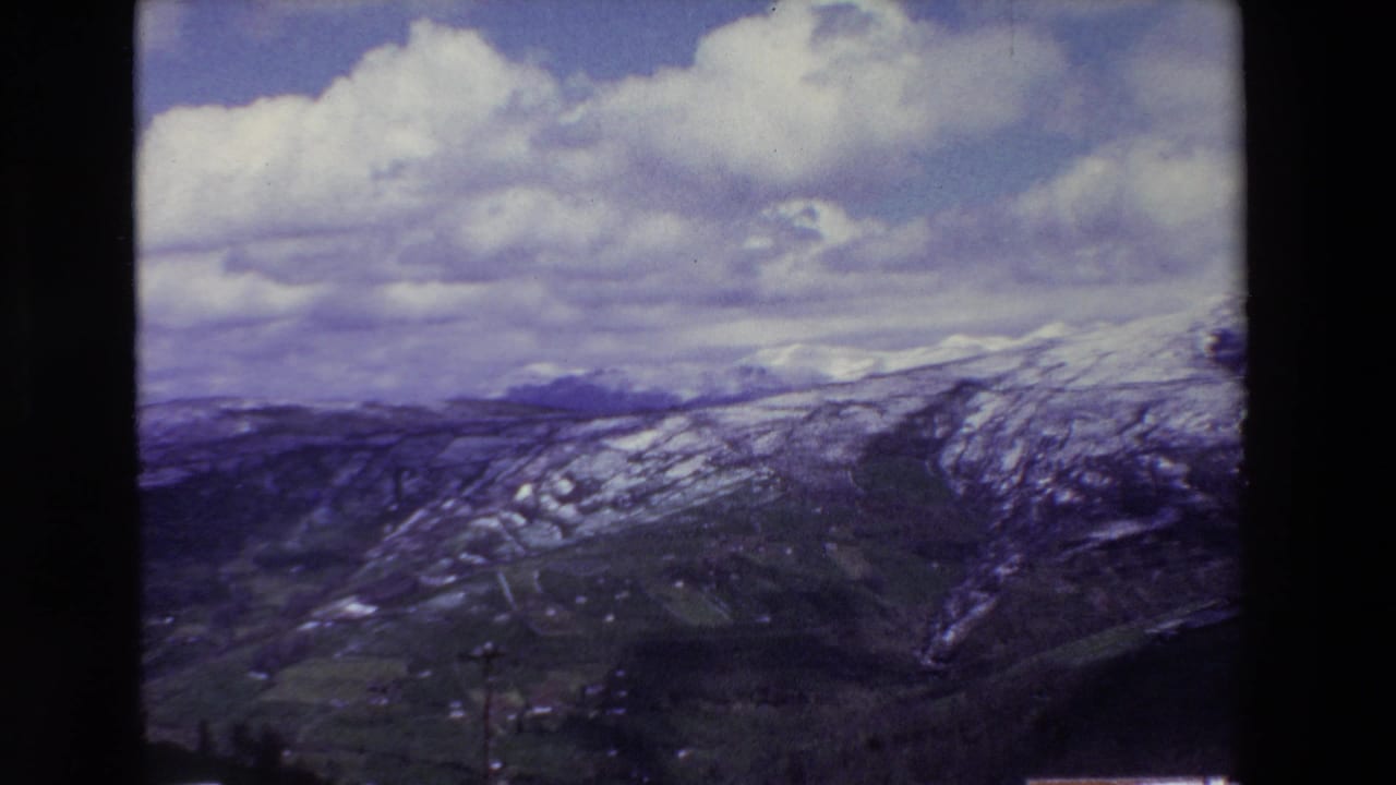 A 1969 8mm home-movie frame shows snow-streaked mountain ridges and a green valley under a sky of large cumulus clouds, with dark film borders visible.