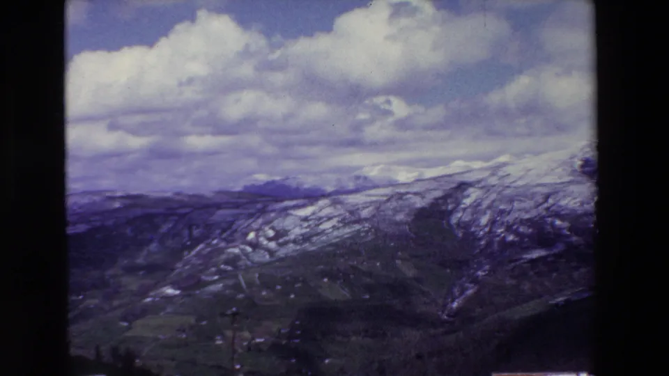 A 1969 8mm home-movie frame shows snow-streaked mountain ridges and a green valley under a sky of large cumulus clouds, with dark film borders visible.
