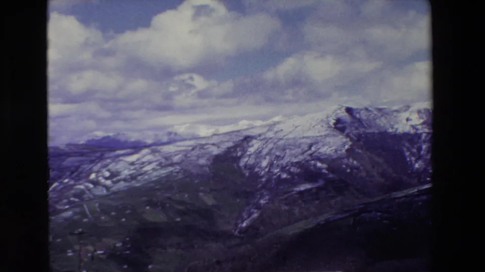 The frame holds on rugged mountain ridges with thin snow bands and darker valley slopes below, retaining the blue-purple cast of the 8mm scan.