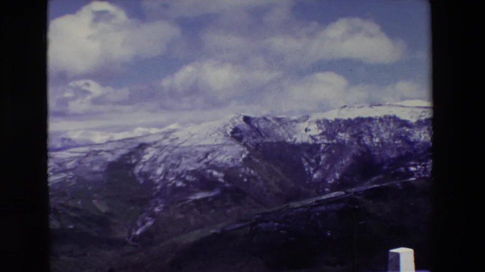 1969 8mm footage of a Spanish mountain landscape with snow on the slopes, a shadowed gorge, blue sky, scattered clouds, and a white roadside post in the foreground.
