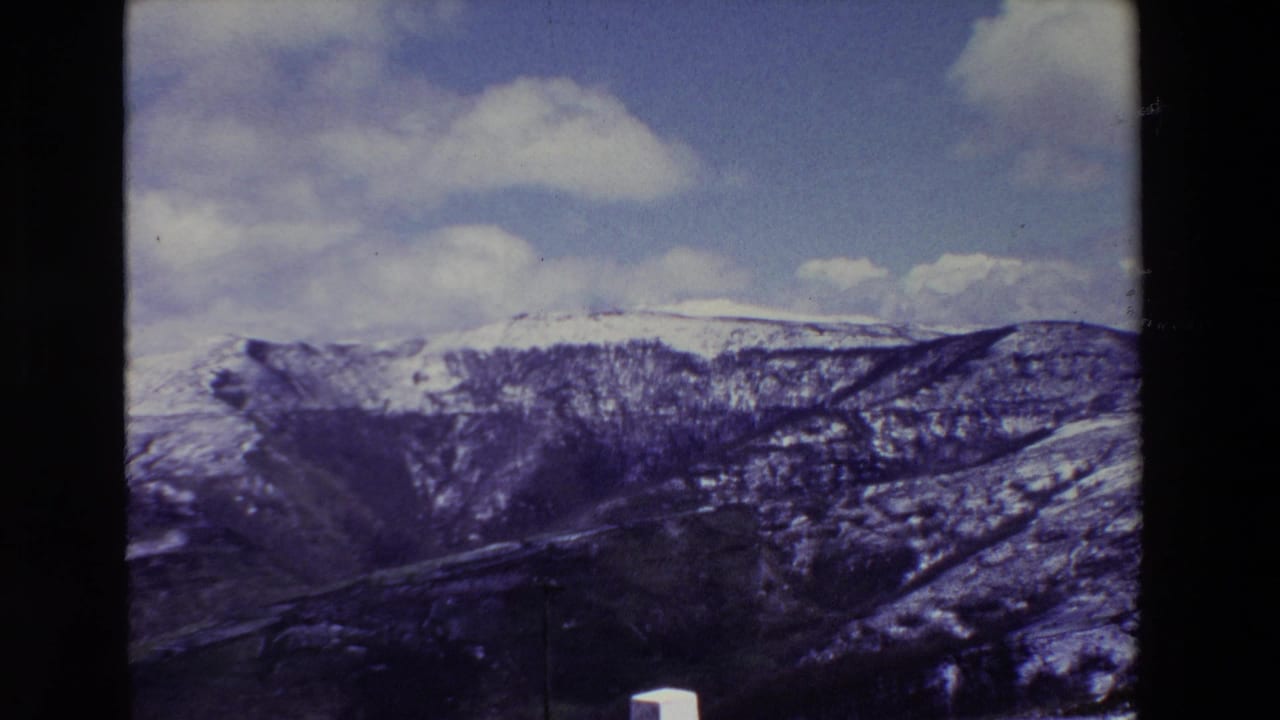 A late-1960s Spanish roadside view shows snow-streaked mountain ridges, a valley or gorge below, scattered white clouds, and a small white post at the lower edge of the frame.