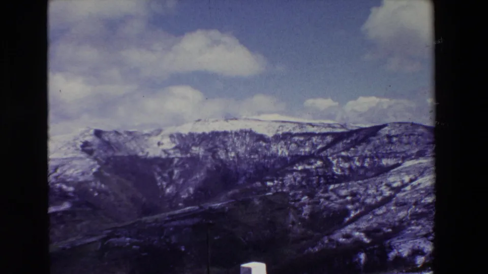 A late-1960s Spanish roadside view shows snow-streaked mountain ridges, a valley or gorge below, scattered white clouds, and a small white post at the lower edge of the frame.