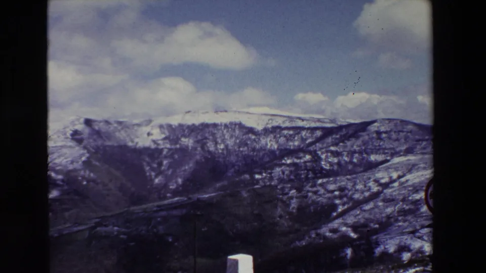 1969 8mm home-movie frame from Spain showing a broad snow-covered mountain ridge, a dark ravine, scattered clouds, and the edge of a circular roadside sign in the foreground.
