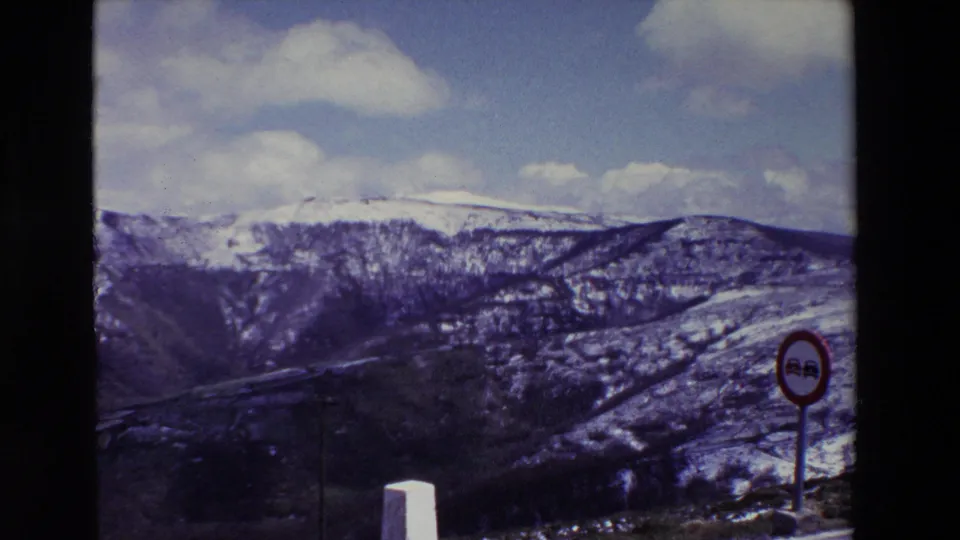 A 1969 Spanish road scene frames snow-covered mountains and a gorge behind a white roadside post and a circular traffic sign, with bright clouds above the ridge.