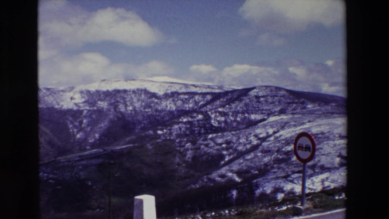 1969 8mm home-movie frame from Spain showing snow-dusted mountain hills, a cloudy sky, a red circular no-passing sign, and a white roadside marker.