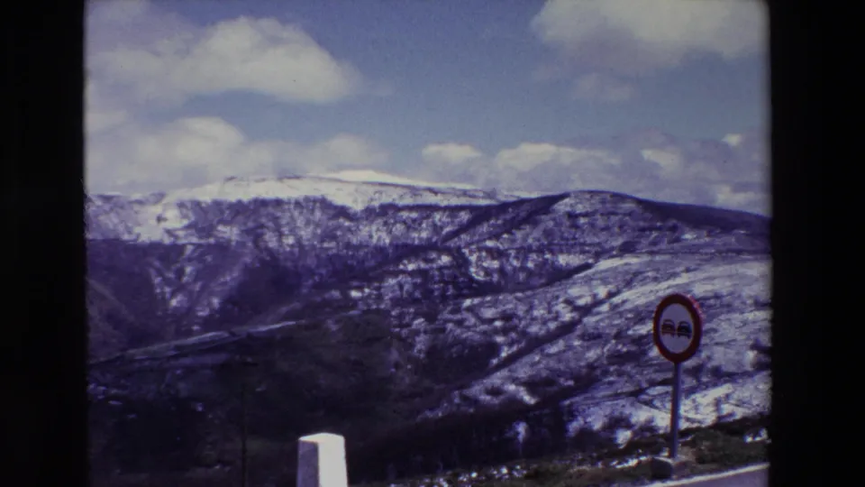 1969 8mm home-movie frame from Spain showing snow-dusted mountain hills, a cloudy sky, a red circular no-passing sign, and a white roadside marker.