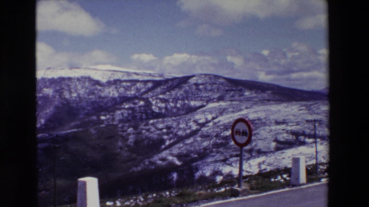1969 8mm frame showing a snowy mountain landscape in Spain with a red roadside no-passing sign, white concrete markers, and a utility pole near the road.