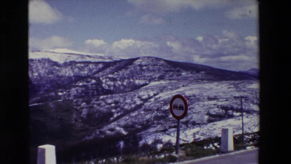 1969 8mm home-movie still from Spain showing a red no-passing sign beside a mountain road, with snow patches across the hills in the background.