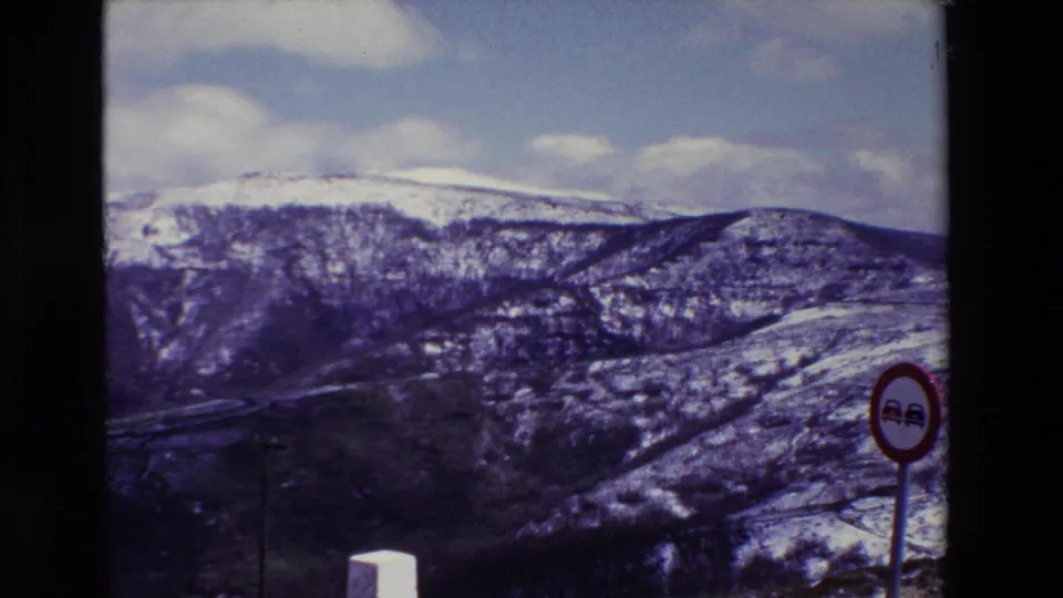1969 8mm frame showing snow-dusted mountains under clouds, with the red no-passing sign partly near the right edge and roadside markers in the foreground.