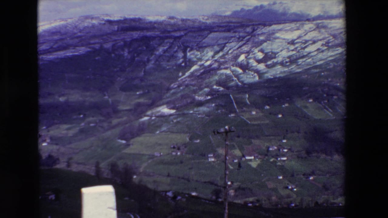 1969 archival 8mm frame showing a snow-dusted rural hillside above green fields, scattered houses, bare trees, and a foreground utility pole, with Spain supplied in the clip metadata.