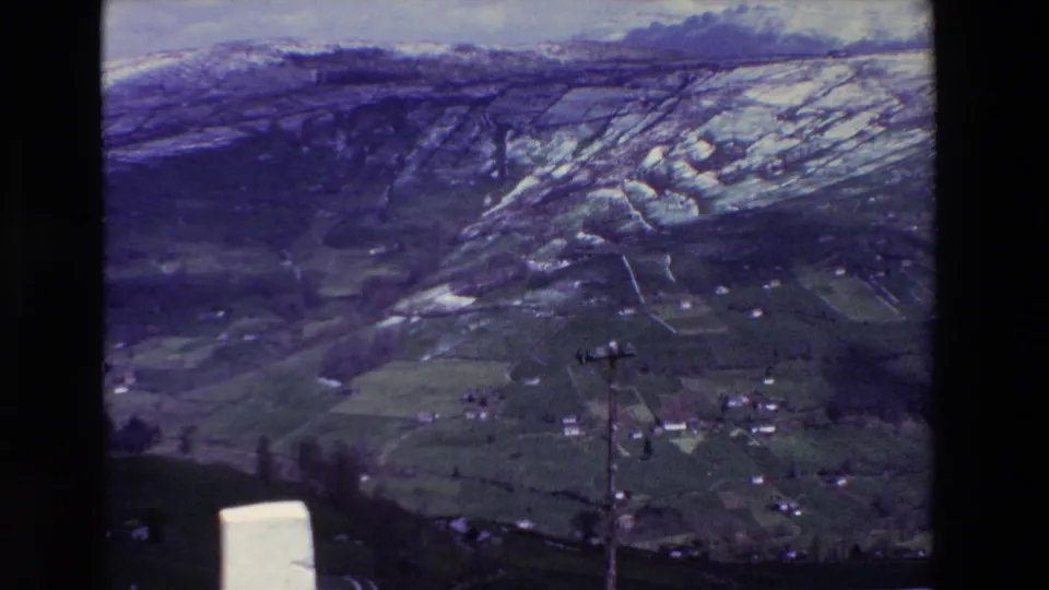 1969 archival 8mm frame showing a snow-dusted rural hillside above green fields, scattered houses, bare trees, and a foreground utility pole, with Spain supplied in the clip metadata.