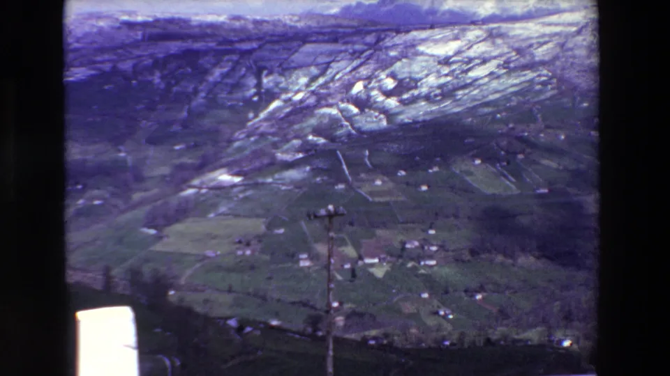 1969 home-movie frame showing a snow-dusted hillside over patchwork fields and small rural houses, with a utility pole and soft purple film color visible.