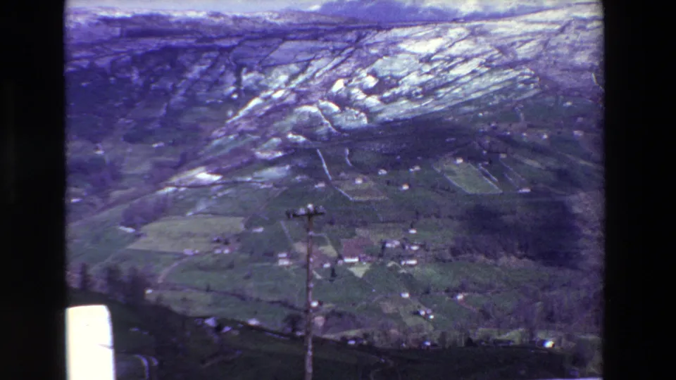 Archival 8mm frame from the supplied Spain 1969 clip showing a wide rural hillside view with snow patches, fields, houses, trees, and visible film gate borders.