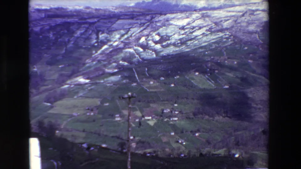 1969 rural Spain aerial 8mm frame showing patchwork fields, scattered pale farm buildings, distant hills, and a wooden utility pole in the foreground.