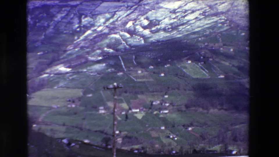 1969 rural Spain aerial frame with farmland divided by pale walls, small white structures, hills beyond the valley, and a wooden utility pole near the lower center.