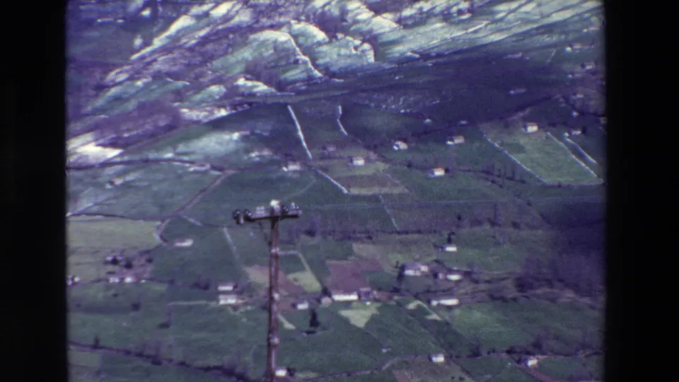1969 rural Spain 8mm frame showing a closer aerial view of green hillside fields, small farm buildings, pale boundary walls, and a foreground utility pole.