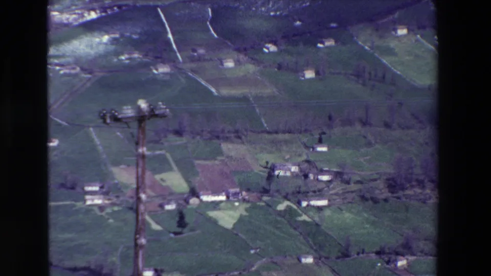 1969 rural Spain aerial frame with divided green fields, scattered light-colored buildings, hillside contours, and a wooden utility pole standing in the foreground.