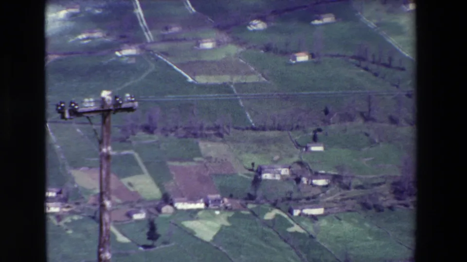 1969 8mm view of green Spanish countryside with scattered farmhouses, field boundaries, and a foreground utility pole.