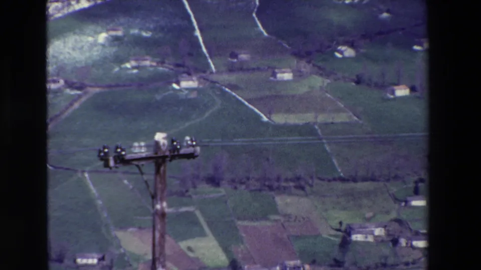 Aerial-style 8mm frame of Spanish farmland, small white buildings, and power lines across rolling green terrain.