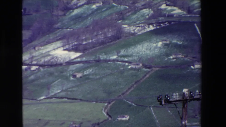 Vintage 8mm countryside view showing rolling Spanish fields, pale ground markings, a small building, and partial utility hardware near the frame edge.
