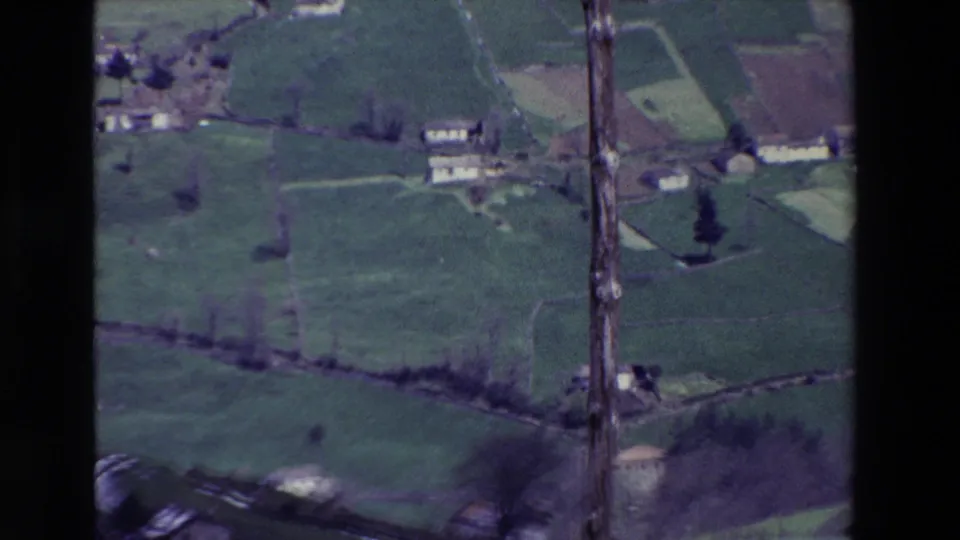 1969 8mm frame of rural Spain with a tall utility pole, overhead wires, green fields, and scattered farm buildings below.