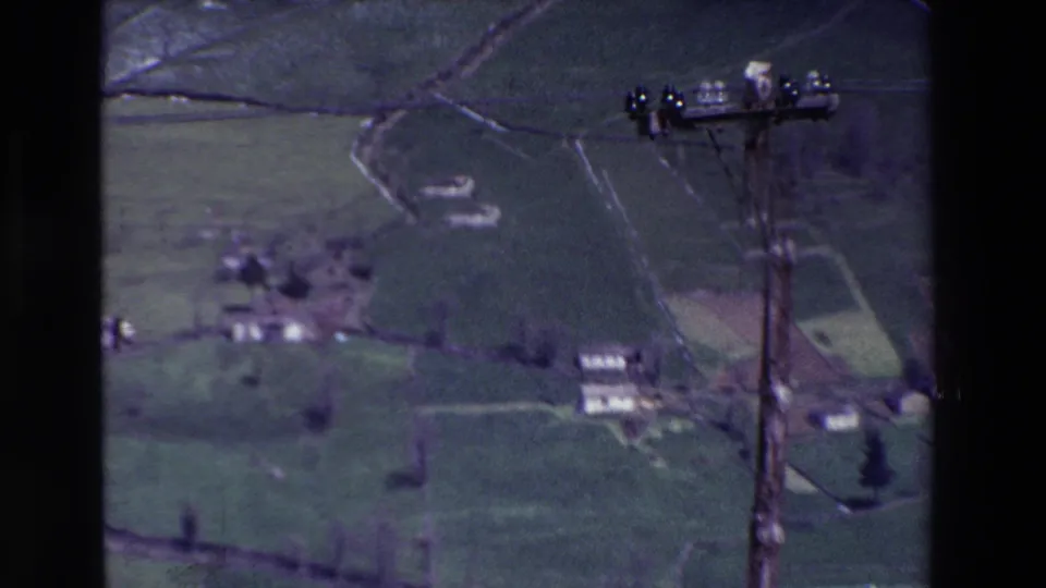 Soft 8mm frame showing rural patchwork fields, small distant houses, hedgerows, and a wooden utility pole with wires at the edge of the view.