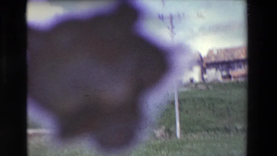 Out-of-focus 8mm frame with a dark foreground blur, grassy countryside, a roadside post, and low buildings in the background.
