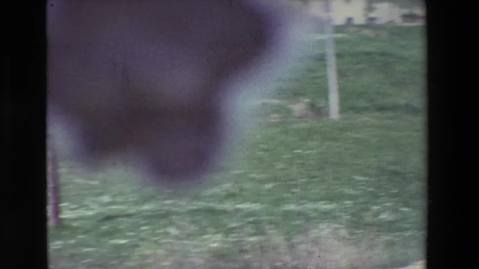Soft, handheld 8mm frame of a grassy rural area with a pale utility pole and a dark out-of-focus shape partly covering the upper left of the view.