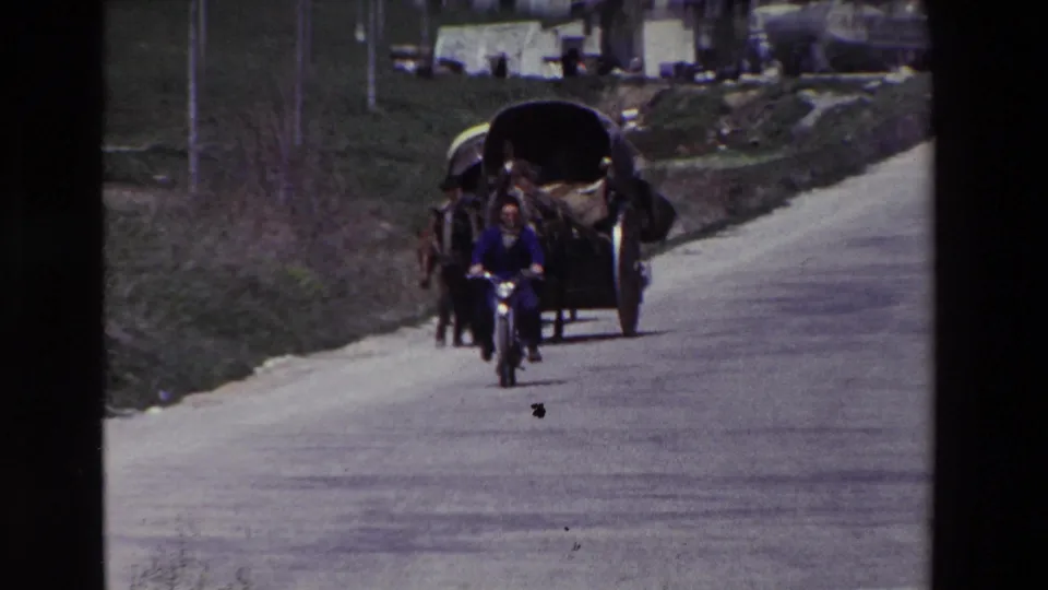 A bicycle rider travels along a paved rural road while a horse-drawn cart, roadside bank, utility poles, and distant buildings appear behind them in the supplied Spain home-movie context.