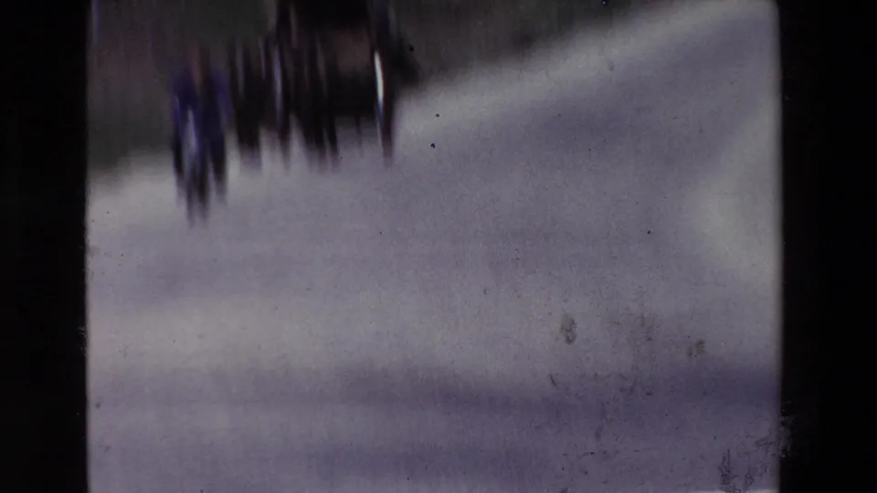 A motion-blurred 8mm frame shows several distant figures or riders on a pale paved road, with the camera movement and film texture obscuring individual details.