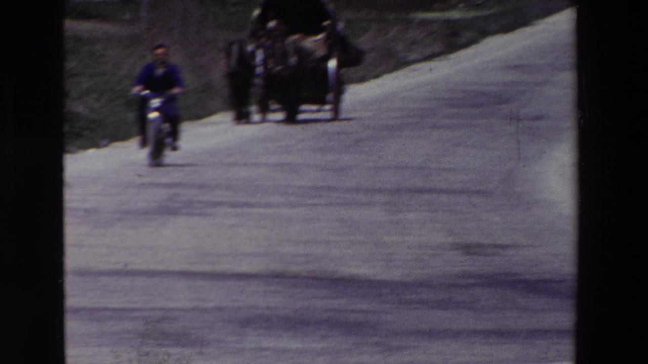 A man on a motorcycle approaches along a pale rural road while a cart or carriage and other figures appear in the distance, consistent with archival home-movie footage from Spain in 1969.